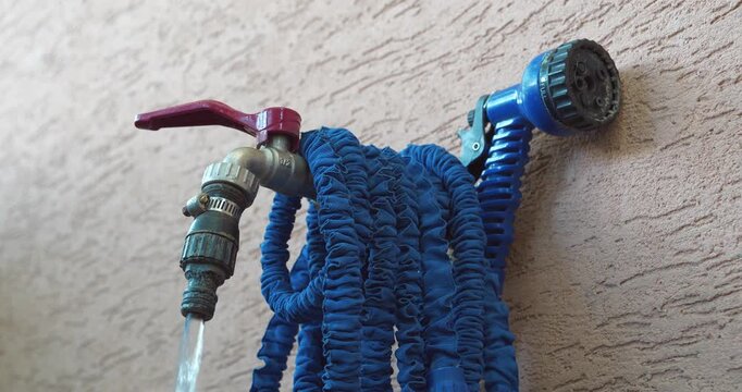 Water tap for watering garden plot outside building. Water is pouring out of tap. Man turns off valve and cut off water. Blue flexible hose with watering nozzle is hanging on tap