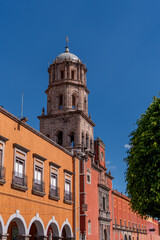 Fototapeta premium A tall tower with a dome sits in front of a row of buildings. The buildings are orange and red. Historic center of Queretaro, colonial architecture, decorations for the celebration of Mexico's Indepen