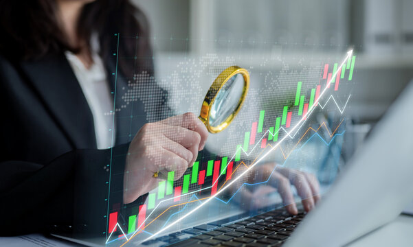 A businesswoman uses a magnifying glass to examine stock market trends on a laptop, symbolizing financial analysis and strategic growth in a modern office environment. Impute - Powered by Adobe