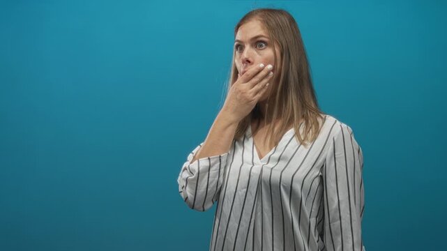 Young blonde woman with finger to lips for silence in blue studio with surprised wide eyed gaze; shock.