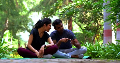 Indian beautiful young couple checking smartphone during yoga break sitting on mat in park outdoor on a sunny day, sharing wellness insights, relaxing together, enjoying peaceful fitness lifestyle - Powered by Adobe