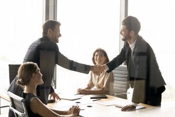 Two happy millennial businessmen shaking hands at round table meeting