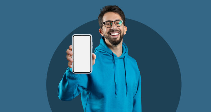 A young man is smiling and showing a smartphone with a blank screen, standing against a simple blue background. He is wearing a bright blue hoodie and shows excitement about technology. - Powered by Adobe
