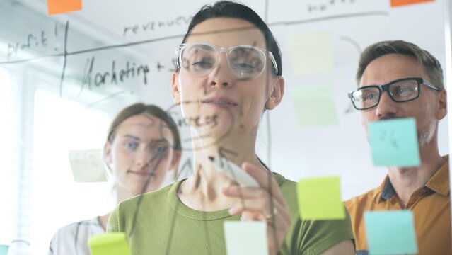 Business people team brainstorming strategic goals, mapping innovative concepts on transparent whiteboard with colorful sticky notes and black marker in modern office - Powered by Adobe