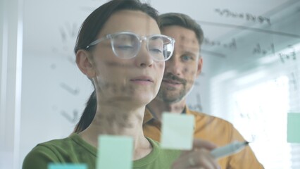 Businesswoman wearing glasses, writing on a glass board and organizing sticky notes, collaborates with adult businessman observing her strategy planning in a bright office