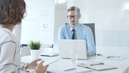 Business colleagues working together at sleek office desk, laptop and tablet open, paperwork spread, conveying professional teamwork and productivity in contemporary workspace