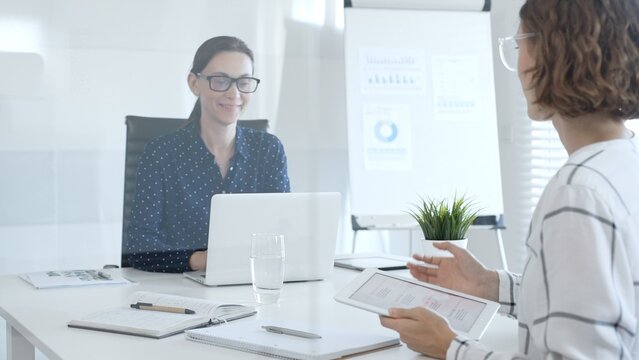 Professional women collaborating at white workspace, reviewing digital content on tablet and laptop during strategic work session