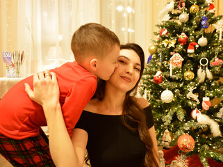 A woman and a boy sitting on the floor in front of a decorated Christmas tree. Garlands, ornaments, and baubles glitter on the tree. The comfort and joy of a family holiday