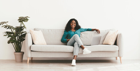 Obraz premium Joyful young african american woman in nice casual outfit relaxing on couch at home, using laptop, watching movie or using dating website, smiling at camera, copy space, full size shot