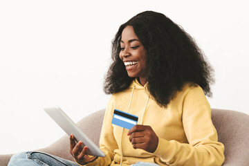 Pretty young black lady relaxing in arm chair with modern digital tablet and credit card, order food or paying for goods and services online, using financial mobile app on pad, copy space