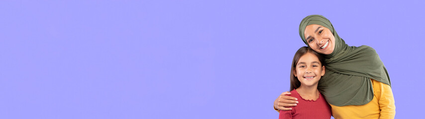 Happy Middle Eastern Mother Embracing Her Daughter Standing On Blue Background In Studio, Smiling To Camera. Arabic Mommy Wearing Hijab Hugging Child Posing Together