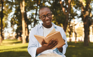 Fototapeta premium Happy african american student guy reading book outdoors, preparing for lectures while sitting on bench in university campus or park, free space