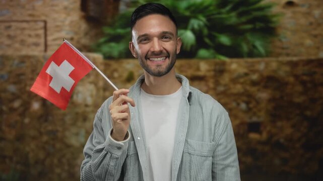 Young man holding switzerland flag smiling in an old town street with urban background outdoors and greenery.
