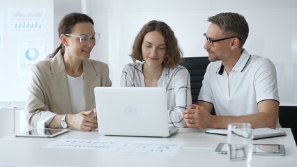 Professional team reviewing project data on laptop, working together in modern office environment, collaborating with focused corporate energy