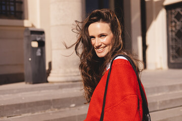 Long brunette hair woman go upstairs, walking on the street. happy smiling woman wear red sweater, black bag. Woman turn her head back, turn around laughing, toothy smile.