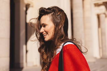 Side view of smiling young woman with long hair wearing a red sweater, walking outdoors on a sunny...