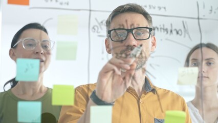 Professional team brainstorming project strategy, placing colorful sticky notes on glass board, analyzing data, sharing insights, developing collaborative solutions