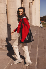 Naklejka premium Photo of lovely positive cute long hair brunette woman going upstairs in old building, she look happy, wear red sweater, beige pants and black bag style fashion outfit. Full length portrait. 