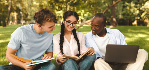 Multiracial students friends learning together, reading books and using laptop computer, sitting on grass in university campus park outdoors