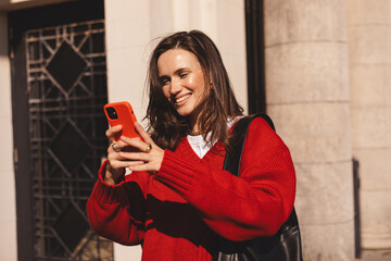 Attractive brunette woman wear red sweater hold mobile phone look happy and smiling. Amazing enjoy woman walk near old building. Portrait of young woman recording voice messages, watch online map.