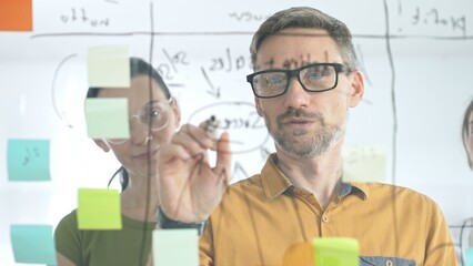 Professional team brainstorming project strategy, placing colorful sticky notes on glass board, analyzing data, sharing insights, developing collaborative solutions