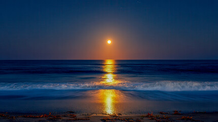 Obraz premium Full moon glowing brilliantly over calm dark ocean waters reflecting golden light onto gentle waves approaching sandy shore at night under clear sky