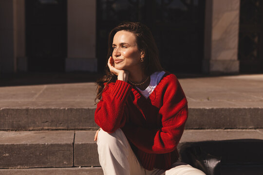 Beautiful smiling fashionable businesswoman sitting on the stairs in front of building in center and posing, laughing. Woman wear red sweater, hold her head.