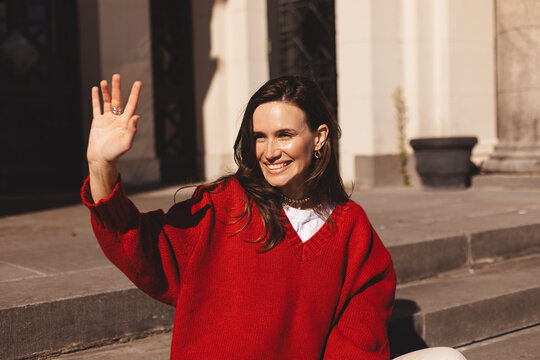 Smiling young brunette woman waving with a friendly cheerful smile. Woman waving her hand standing sitting on the stairs. Girl wear red sweater meeting friends, hi, bye. - Powered by Adobe