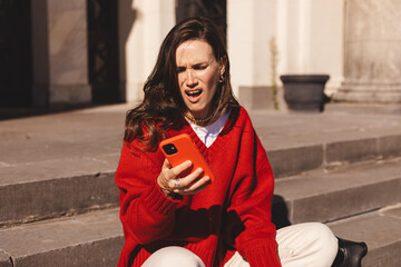 Beautiful upset woman sitting outdoor on the stairs. Sad angry female look angry, mad, holding mobile phone, yelling. Screaming woman looking phone feels shock upset worries bad news problems.