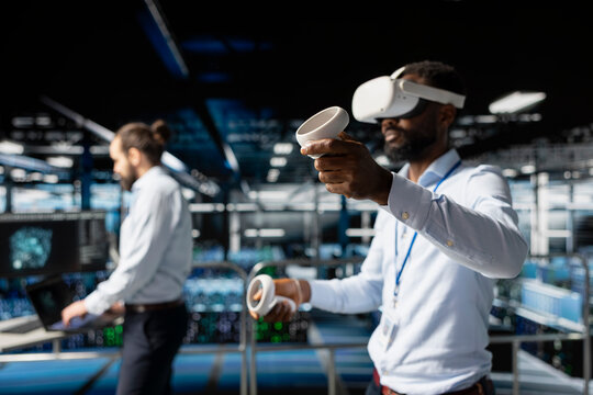 African american IT professional using VR technology to simulate AI neural network systems. Standing on industrial platform in data center using virtual reality headset with futuristic tools.
