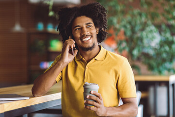 Coffee Break In Cafe. Happy Black Guy Holding Cup With Takeaway Drink And Talking On Cellphone While Sitting At Table In Cafe, Cheerful Handsome African American Man Relaxing In Cozy Cafeteria