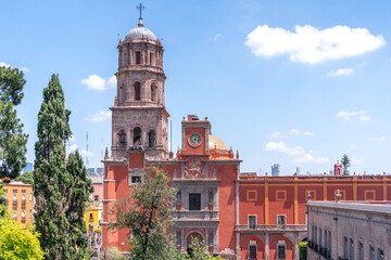 A large red building with a clock tower and a cross on top. The building is surrounded by trees. Historic center of Queretaro, colonial architecture, decorations for the celebration of Mexico's Indepe