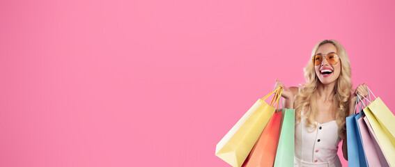 Happy Customer. Portrait of happy emotional young woman wearing sunglasses holding colorful shopping bags with gifts, cheerful lady looking aside at free copy space, pink studio background, banner