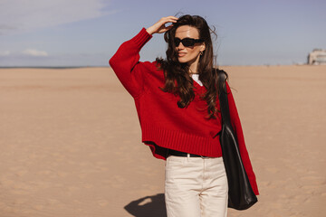 Long brunette hair woman walking outdoor on the beach coast, she wear red sweater and black bag, eyeglasses on shoulder, sensual smile woman fix her hear, spring, autumn season.