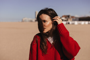 Fototapeta premium Long brunette hair woman walking outdoor on the beach coast, she wear red sweater and black bag on shoulder, sensual calm woman fix her hair, spring, autumn season. Enjoy of sunny day.