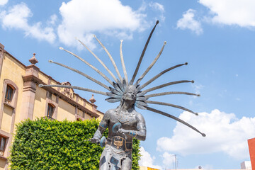 A statue of a man with a feather headdress. The statue is surrounded by green bushes. Historic center of Queretaro, colonial architecture, decorations for the celebration of Mexico's Independence Day.