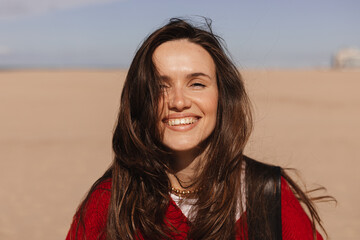Happy long brunette hair woman wear red sweater, black bag while walking outdoor on beach coast,...
