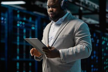 Server room IT expert listening music while fixing network issues affecting rigs performance. Data center programmer wearing audio headphones running system diagnostic tests on tablet