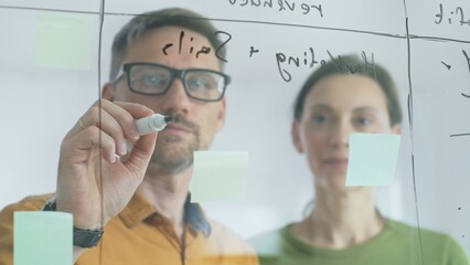Professional team members brainstorming strategy, writing notes on transparent whiteboard with colorful sticky papers during collaborative meeting in office. Business people at work