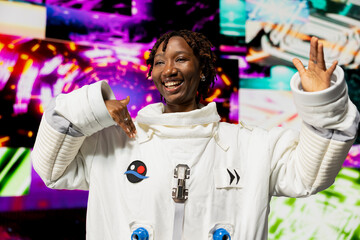 Fototapeta premium Joyous woman wearing astronaut suit doing dance moves with hands reacting to abstract colorful visuals on digital background. Upbeat cosmonaut having fun, isolated against bright neon vortex