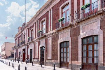 Naklejka premium A row of red brick buildings with balconies and potted plants. The buildings are old. Historic center of Queretaro, colonial architecture, decorations for the celebration of Mexico's Independence Day.