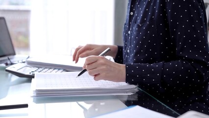 Businesswoman making notes while reviewing ring folder of financial documents with magnifying glass at desk in modern office. Audit and taxes in business