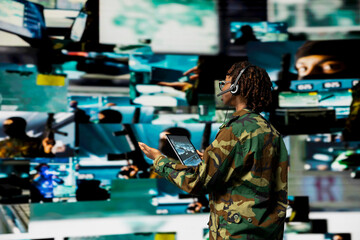 Army telecommunications operator uses tablet and headset to manage secure military communications. Soldier coordinates mission support from command center with surveillance screens in background