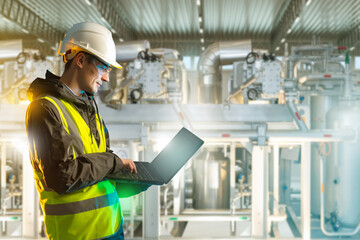 Engineer in helmet and safety vest uses laptop while inspecting industrial equipment and pipelines inside factory. Industry, Engineering, Manufacturing.