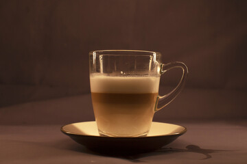 A transparent glass cup filled with beautifully layered coffee sits elegantly on a saucer against a neutral background