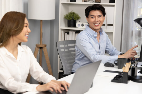 Smiling Asian man talking with woman colleague in planning marketing report project on laptop casual day while searching database on working desk at modern home office at side view image. Infobahn.