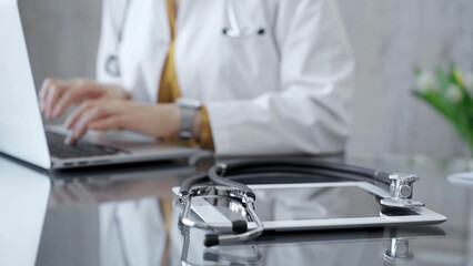 Close-up of doctor's desk with stethoscope and tablet computer. Physician is using a laptop at the background. Medicine concept