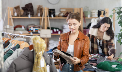 Young woman standing in store and choosing gloves in front of Armenian woman choosing jersey sweaters