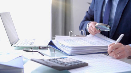 Obraz premium Businessman using a calculator and magnifying glass while analyzing financial documents at desk in fair modern office. Audit and taxes in business