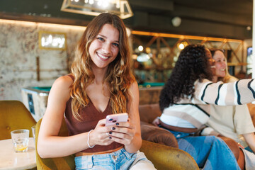 Young woman looking at camera while friends take a selfie, enjoying time in a bar